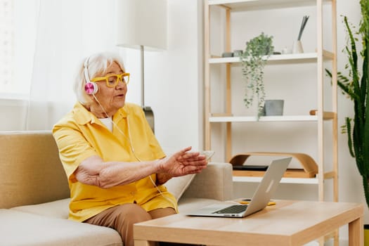 Elderly woman wearing headphones with a laptop communication online video call smile happiness, sitting on the couch at home and working in a yellow shirt, the lifestyle of a retired woman. High quality photo