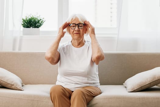 Elderly woman severe pain in the head sitting on the couch, health problems in old age, poor quality of life. Grandmother with gray hair holding her head, migraine and high blood pressure. High quality photo