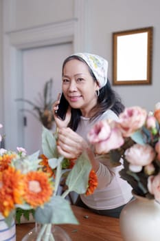 Startup, small business, flower shop. Older florist putting a flower on a vase and talking with customer by phone.