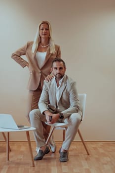 A business couple poses for a photograph together against a beige backdrop, capturing their professional partnership and creating a timeless image of unity and success