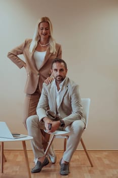 A business couple poses for a photograph together against a beige backdrop, capturing their professional partnership and creating a timeless image of unity and success