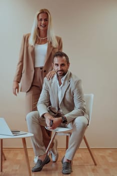 A business couple poses for a photograph together against a beige backdrop, capturing their professional partnership and creating a timeless image of unity and success