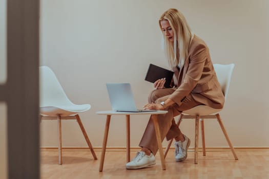 A professional businesswoman sits on a chair, surrounded by a serene beige background, diligently working on her laptop, showcasing dedication and focus in her pursuit of success.