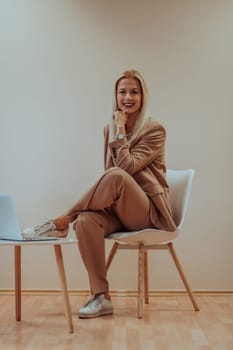 A professional businesswoman sits on a chair, surrounded by a serene beige background, diligently working on her laptop, showcasing dedication and focus in her pursuit of success.