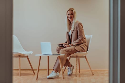 A professional businesswoman sits on a chair, surrounded by a serene beige background, diligently working on her laptop, showcasing dedication and focus in her pursuit of success.