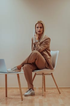 A professional businesswoman sits on a chair, surrounded by a serene beige background, diligently working on her laptop, showcasing dedication and focus in her pursuit of success.