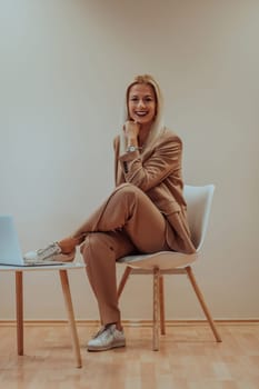 A professional businesswoman sits on a chair, surrounded by a serene beige background, diligently working on her laptop, showcasing dedication and focus in her pursuit of success.