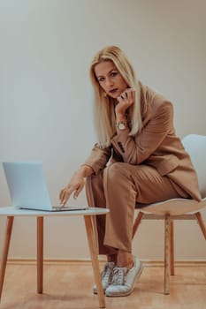 A professional businesswoman sits on a chair, surrounded by a serene beige background, diligently working on her laptop, showcasing dedication and focus in her pursuit of success.