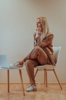 A professional businesswoman sits on a chair, surrounded by a serene beige background, diligently working on her laptop, showcasing dedication and focus in her pursuit of success.