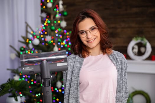 A smiling young woman holds a vacuum cleaner in her hand and a Christmas tree and fireplace can be seen in the background. Domestic cleaning work before Christmas.