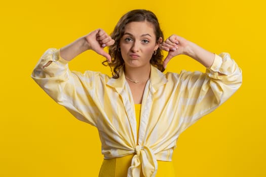 Dislike. Upset unhappy young woman showing thumbs down sign gesture, expressing discontent, disapproval, dissatisfied, dislike. Pretty attractive girl. Indoors isolated on yellow studio background