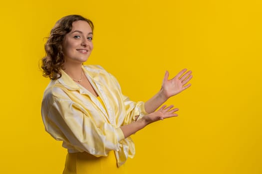Young woman showing thumbs up and pointing right empty place, advertising area for commercial text, copy space for goods promotion advertisement. Girl isolated on yellow studio background. Lifestyles