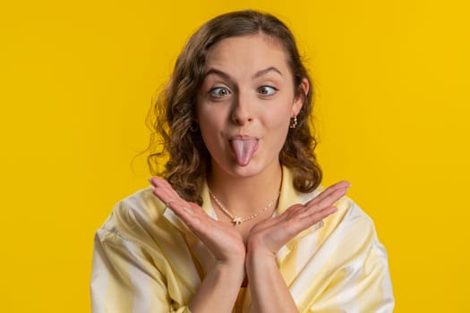 Funny comical playful young woman making silly facial expressions and grimacing, fooling around, showing tongue, idiotic expression. Girl isolated alone on yellow studio background. People lifestyles