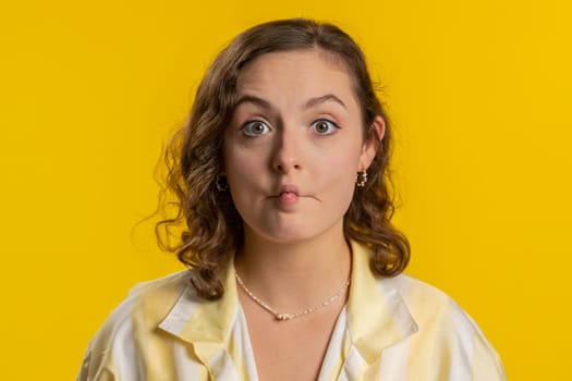 Surprised funny comical playful young woman making fish grimace pout lips, smooching choking, looking with shocked dumb idiotic expression, doing duck face, fooling around. Girl on yellow background