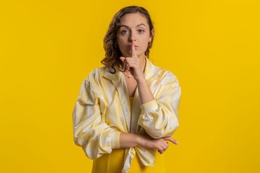 Shh be quiet please. Young woman presses index finger to lips makes silence gesture sign do not tells secret, stop gossip. Pretty attractive brunette girl isolated alone on yellow studio background