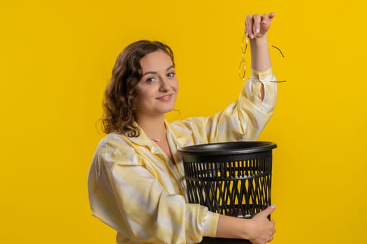 Brunette attractive woman taking off throwing out glasses into bin after medical vision laser treatment therapy surgery looking smiling at camera. Pretty girl isolated on yellow studio background