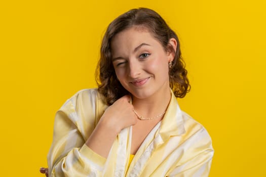 Smiling playful happy young woman blinking eye, looking at camera with smile, winking and flirting, expressing optimism. Excited girl isolated alone on yellow studio background. Lifestyles