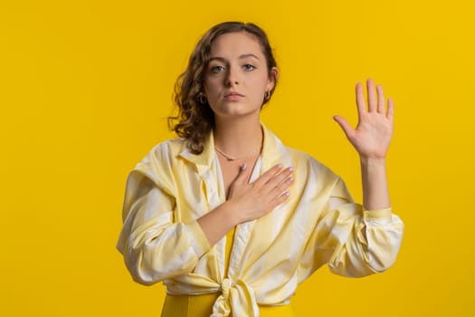 I swear to be honest. Sincere responsible young woman raising hand to take oath, promising to be honest and to tell truth, keeping hand on chest. Girl isolated on yellow studio background. Lifestyles
