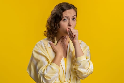 Unhealthy young woman coughing covering mouth with hand, feeling sick, allergy or viral infection symptoms, fever pain virus. Pretty attractive brunette girl isolated alone on yellow studio background