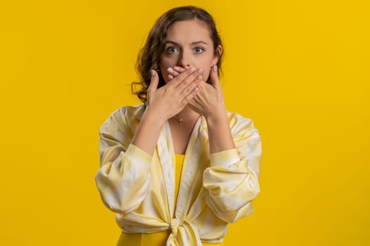 I will not say anything. Frightened woman closing mouth with hands, looking intimidated scared at camera, gestures no, refusing to tell terrible secret, unbelievable truth. Girl on yellow background
