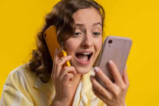 Irritated nervous stressed young woman talking screaming on two mobile phones having conversation conflict quarrel complaint dispute discuss solve problem. Girl isolated on yellow studio background