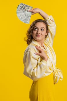 Rich pleased boss young woman waving money dollar cash banknotes bills like a fan, success business career, lottery winner, big income, wealth. Brunette girl isolated on yellow background, indoors