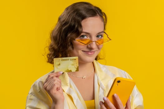 Sincere young woman customer using credit bank card and smartphone while transferring money, purchases online shopping, payment. Finance and internet. Pretty girl isolated alone on yellow background