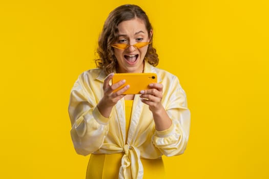 Worried funny addicted woman enthusiastically playing racing video games on mobile phone. Happy pretty young girl using smartphone gadget app with drive simulator isolated on yellow studio background