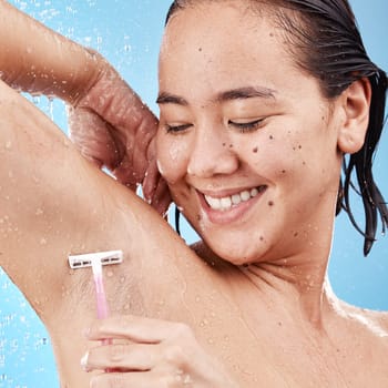 Shower, woman shave armpit with razor and clean beauty with water and hygiene for grooming against blue studio background. Happy Asian model, healthy skin and wet with hair removal and shaving