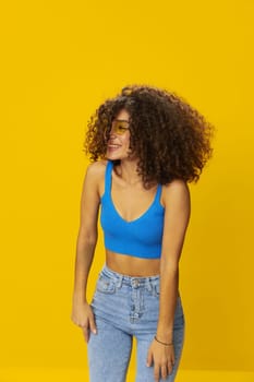 Woman with curly afro hair in a blue T-shirt on a yellow background dancing flying hair with sunglasses yellow, hand signs, look into the camera, smile with teeth and happiness, copy space. High quality photo