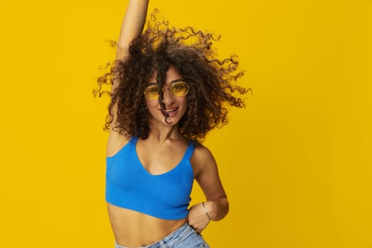 Woman with curly afro hair in a blue T-shirt on a yellow background dancing flying hair with sunglasses yellow, hand signs, look into the camera, smile with teeth and happiness, copy space. High quality photo