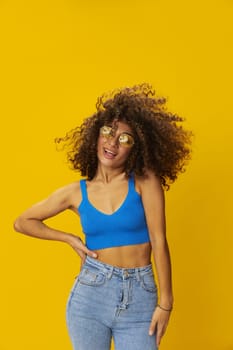 Woman with curly afro hair in a blue T-shirt on a yellow background dancing flying hair with sunglasses yellow, hand signs, look into the camera, smile with teeth and happiness, copy space. High quality photo