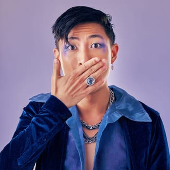 Surprised, shocked and gay man with makeup in a studio for pride, beauty and gender fluid cosmetics. Lgbtq, queer and homosexual guy with wow, surprise or omg expression isolated by purple background.
