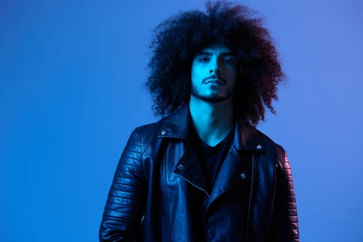 Portrait of fashion man with curly hair on blue background multinational, colored light, black leather jacket trend, modern concept. High quality photo