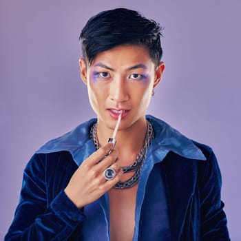 Fashion, beauty and makeup with a man model in studio on a purple background applying red lipstick. Face, portrait and cosmetics with a young asian male indoors to promote a cosmetic product.
