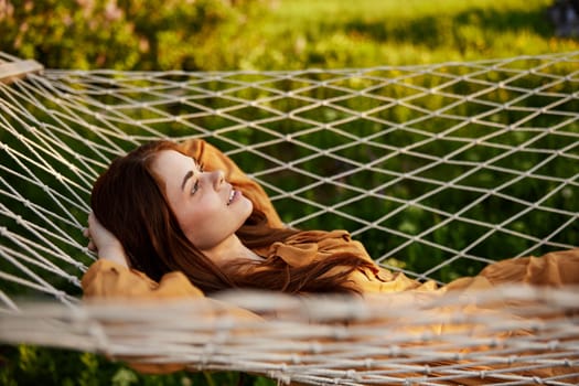 a woman is resting lying in a mesh hammock with her hands behind her head, smiling happily, enjoying a warm day in the rays of the setting sun, lying in an orange dress. High quality photo