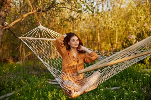 a joyful woman is sitting in a mesh hammock in nature, relaxing and enjoying the rays of the setting sun, straightening her hair with her hands, closing her eyes on a warm summer day. Horizontal photo on the theme of outdoor recreation. High quality photo