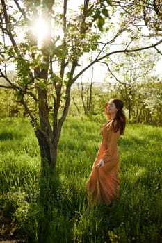 a beautiful, joyful woman stands in a long orange dress, in the countryside, near a tree blooming with white flowers, during sunset, illuminated from behind and touches her long hair. High quality photo