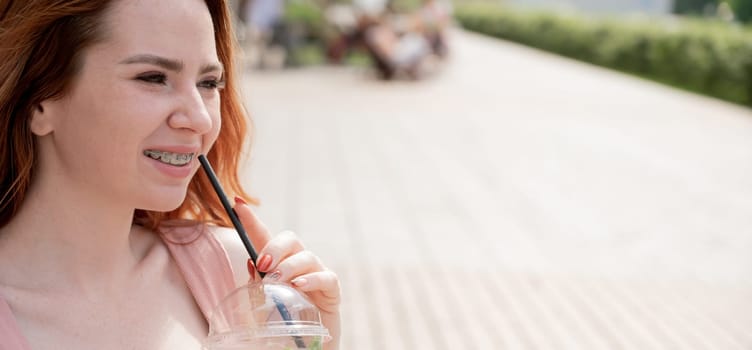 Young beautiful red-haired woman with braces drinks cooling lemonade outdoors in summer. Portrait of a smiling girl with freckles