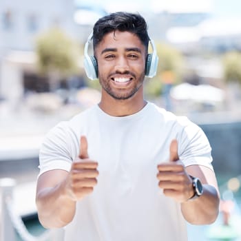 Happy man, headphones and listening to music with thumbs up for winning, fitness or success in city. Portrait of male person, athlete or runner with headset, thumb emoji or yes sign for audio track.