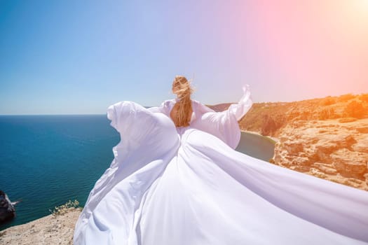 woman sea white dress. Blonde with long hair on a sunny seashore in a white flowing dress, rear view, silk fabric waving in the wind. Against the backdrop of the blue sky and mountains on the seashore