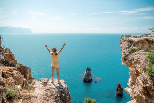 Happy girl stands on a rock high above the sea, wearing a yellow jumpsuit and sporting braided hair, depicting the idea of a summer vacation by the sea