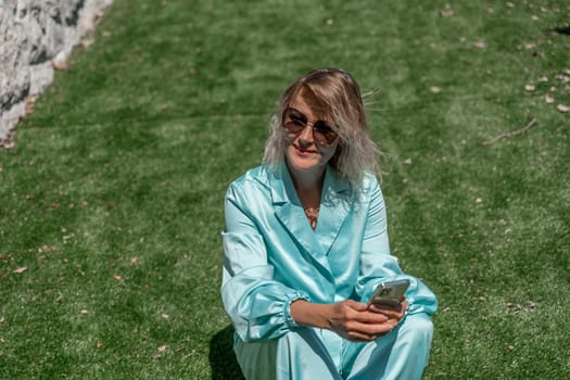 Woman grass. Happy woman with blond curly hair in glasses and in a blue overalls sits on the summer fresh green grass with a phone in her hands