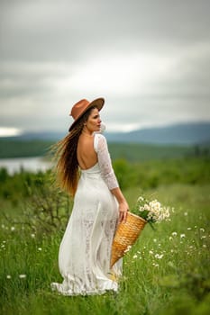 A middle-aged woman in a white dress and brown hat stands on a green field and holds a basket in her hands with a large bouquet of daisies. In the background there are mountains and a lake