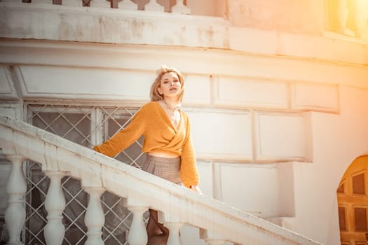 A middle-aged woman looks like a good blonde with curly beautiful hair and makeup on the background of the building. She is wearing a yellow sweater