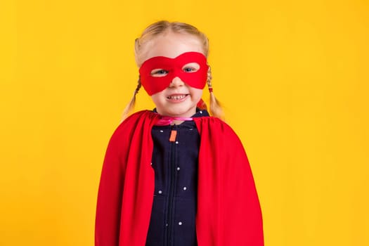 Funny little power superhero child girl in a red raincoat and a mask on a yellow background. Studio photography. Superhero concept.