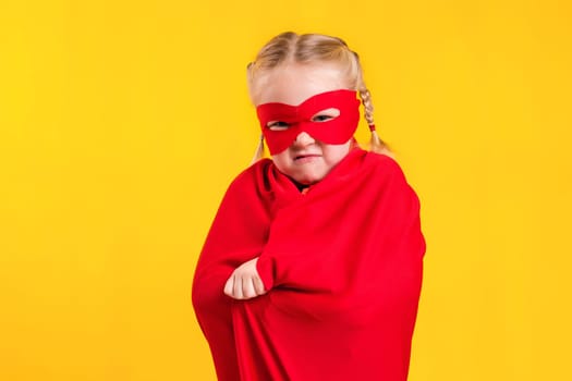 Funny little power superhero child girl in a red raincoat and a mask on a yellow background. Studio photography. Superhero concept.