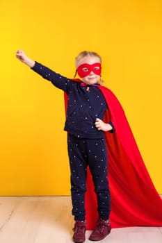 Funny little power superhero child girl in a red raincoat and a mask on a yellow background. Studio photography. Superhero concept.