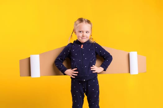 A little child girl is wearing homemade cardboard flying wings, pretending to be a pilot for a craft, imagination or exploration concept. Studio photography on a yellow background