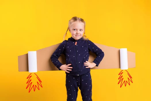 A little child girl is wearing homemade cardboard flying wings, pretending to be a pilot for a craft, imagination or exploration concept. Studio photography on a yellow background